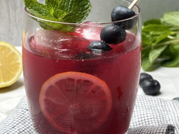 Glass with blueberry ginger lemonade garnished with fresh mint, lemon slice, and berries.