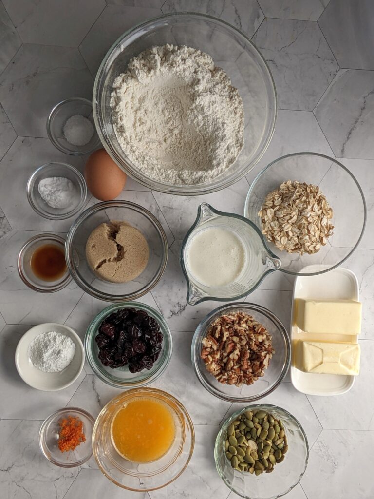 overhead shot of ingredients: flour, butter, rolled oats, orange juice, baking powder and soda, egg, dried cranberries, cinnamon, salt, brown sugar, vanilla, orange zest, cream, pecans, pepitas, confectioner's sugar