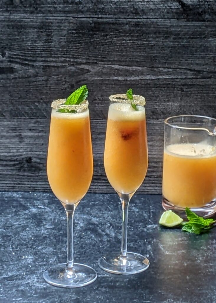 finished frozen peach bellinis with mint garnish and strawberry slices being served in two champagne flutes rimmed with raw sugar