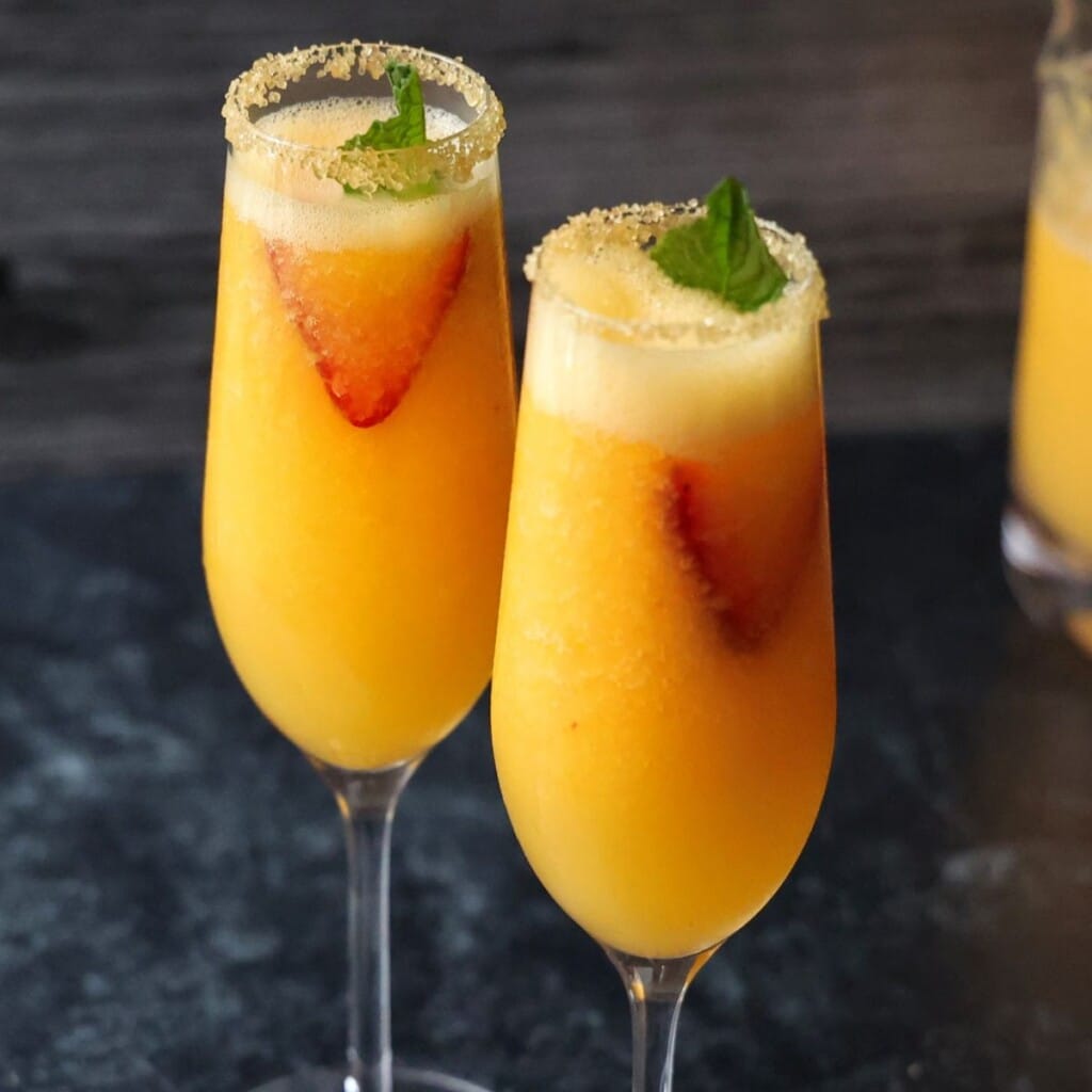 Two frozen peach bellinis in champagne flutes garnished with strawberries and mint.