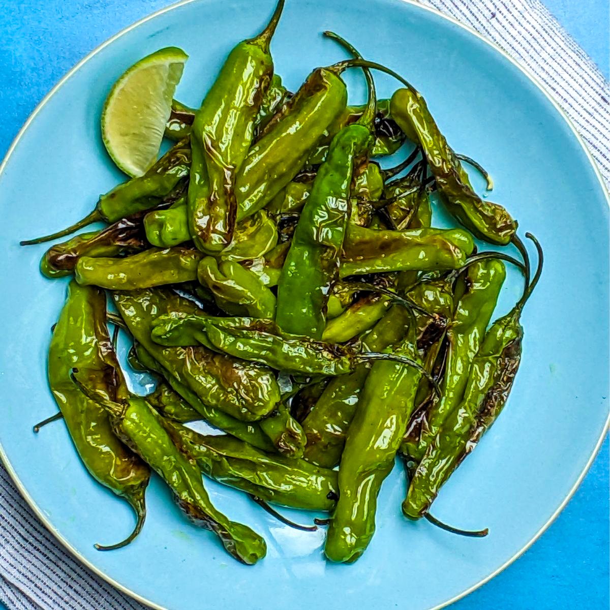 Overhead shot of blue plate with blistered shishitos peppers and wedge of lime.