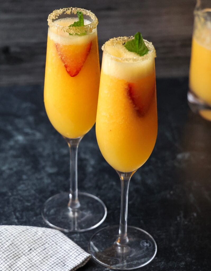 two Frozen Peach Bellinis in sugar rimmed champagne flutes, garnished with a mint leaf and strawberry slicesin
