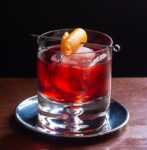 Close up of mezcal negroni in glass with orange twist. The drink is in a dark luxurious bar with a black background.