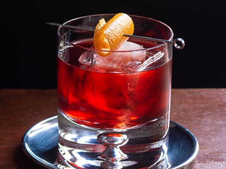 Close up of mezcal negroni in glass with orange twist. The drink is in a dark luxurious bar with a black background.