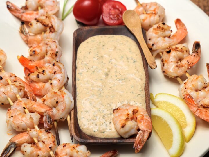 Platter with grilled shrimp on skewers with wooden bowl of remoulade sauce, lemon wedges and garnished with parsley and cherry tomatoes.