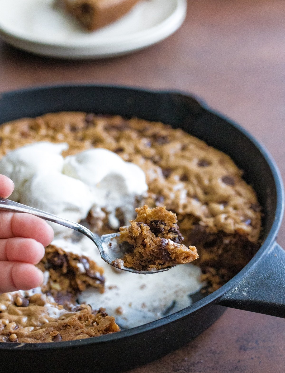 Chocolate chip skillet cookie in a cast iron skillet with scoops of vanilla cream on top and scooping some out with a spoon.