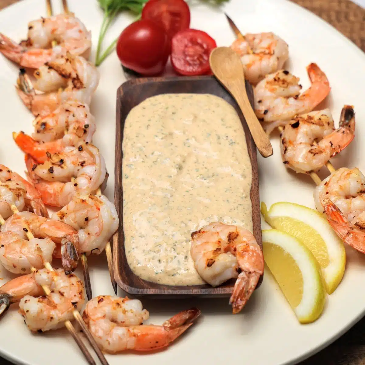 Platter with grilled shrimp on skewers with wooden bowl of remoulade sauce, lemon wedges and garnished with parsley and cherry tomatoes.