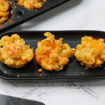 Three Mac and Cheese Bites on a small black rectangular platter with the filled muffin pan in background.