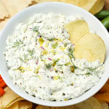 White bowl filled with pickle dip with potato chips in dip and around bowl.