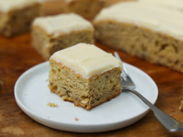 Close up of square of banana bars with cream cheese icing on a white plate with a fork.